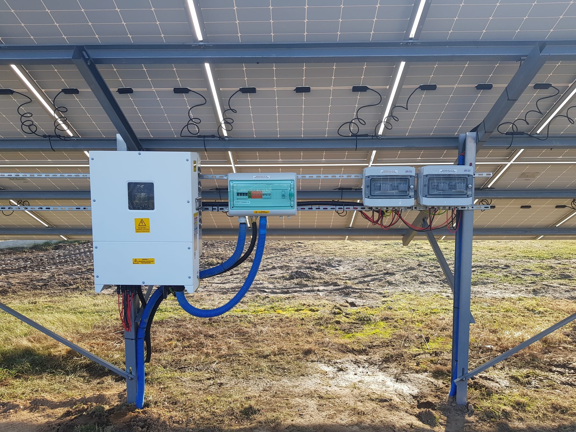 Instalacja paneli słonecznych we Wrocławiu: widok od spodu na konstrukcję, inwerter i skrzynki zabezpieczające, z przewodami w niebieskich peszlach.