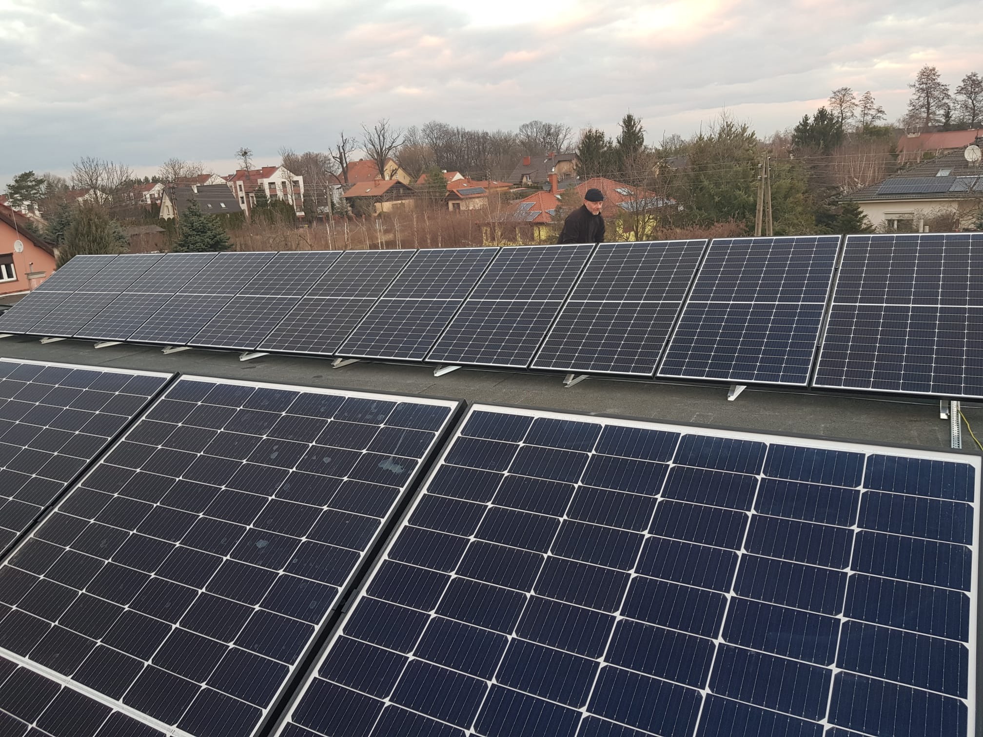 Instalacja paneli słonecznych na dachu domu jednorodzinnego, widok z góry, pracownik w czarnej kurtce i czapce na dachu, w tle zabudowa mieszkalna i pochmurne niebo.