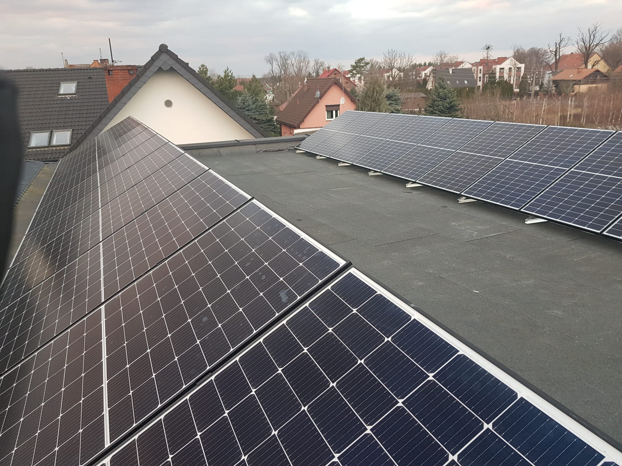 Panele słoneczne zamontowane na płaskim dachu budynku mieszkalnego, widok z góry, pochmurne niebo w tle.