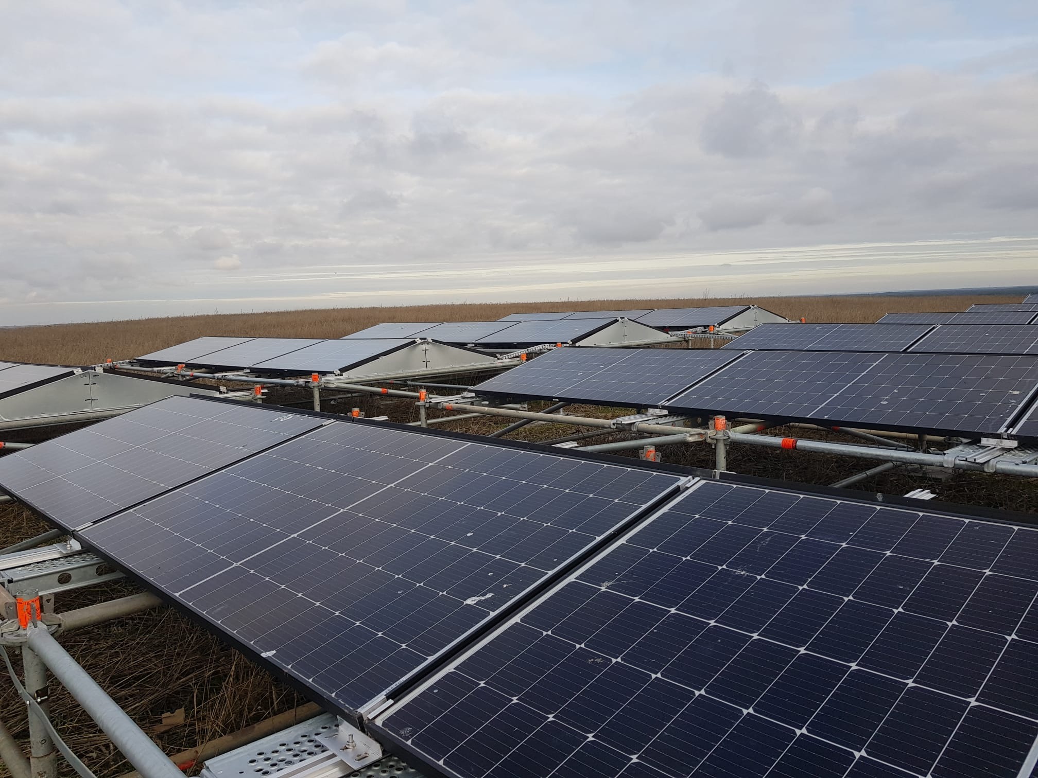 Panel słoneczny zamontowany na metalowej konstrukcji, widok na pole i pochmurne niebo w tle.