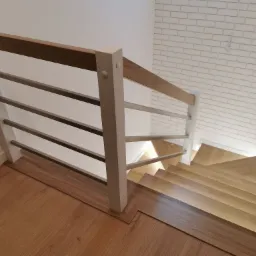 Nowoczesne schody z drewnianymi stopniami i metalowymi relingami, białe słupki, oświetlenie LED przy ścianie, widok z góry na dół.