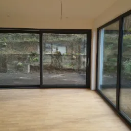 Wnętrze pokoju z nowo ułożonymi panelami podłogowymi w odcieniach jasnego drewna i dużymi przesuwnymi oknami z widokiem na ziemny nasyp porośnięty roślinnością.