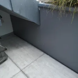 Szara ściana z roślinami na dachu, widoczne łączenia płyt i betonowa podłoga z fugami wypełnionymi brudem.