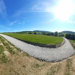 Nowo utwardzona droga z jasnego kruszywa wijąca się przez zielone wzgórze w słoneczny dzień. Widoczne ślady po pracach ziemnych i krajobraz z zabudowaniami w oddali.