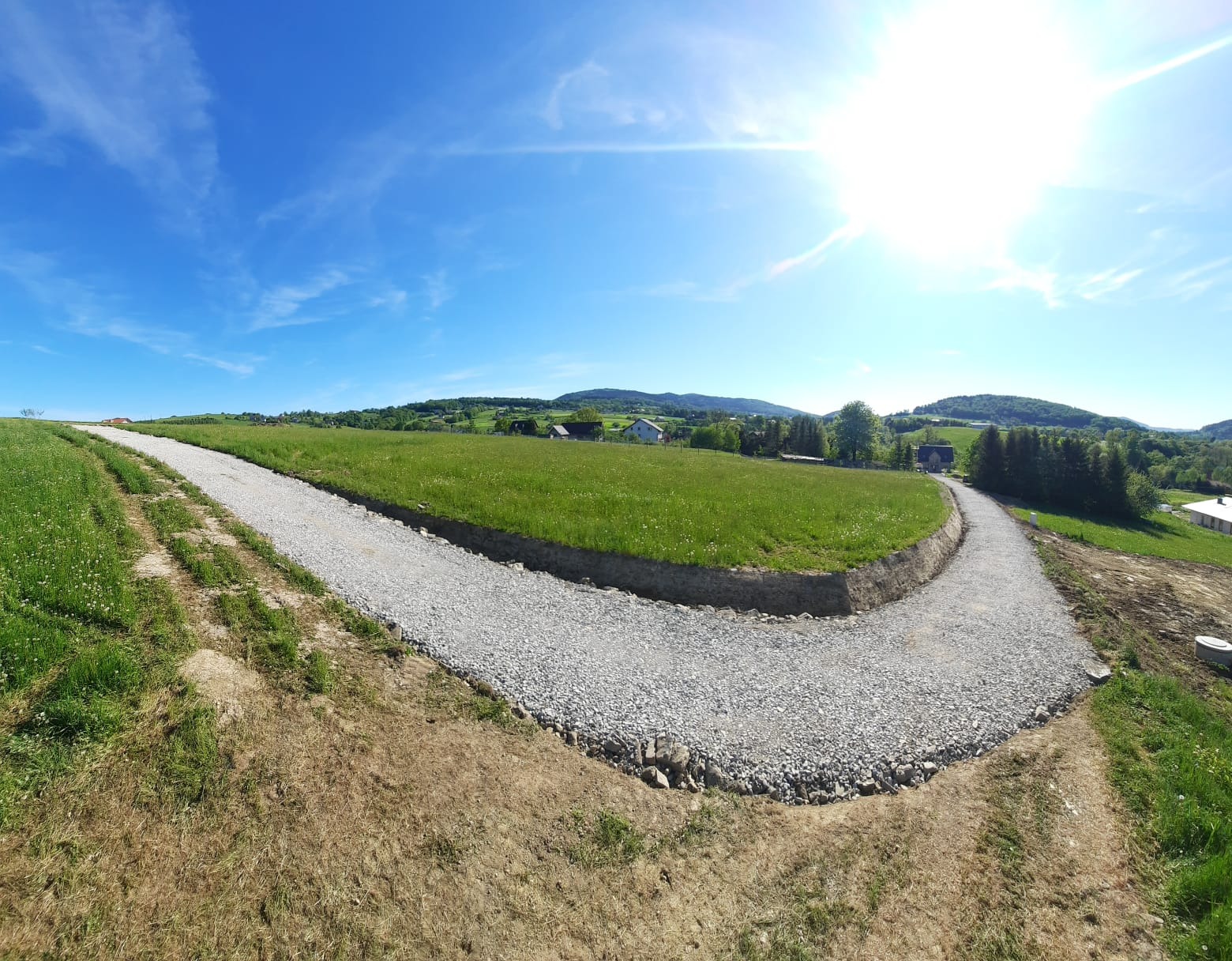 Nowo utwardzona droga z jasnego kruszywa wijąca się przez zielone wzgórze w słoneczny dzień. Widoczne ślady po pracach ziemnych i krajobraz z zabudowaniami w oddali.