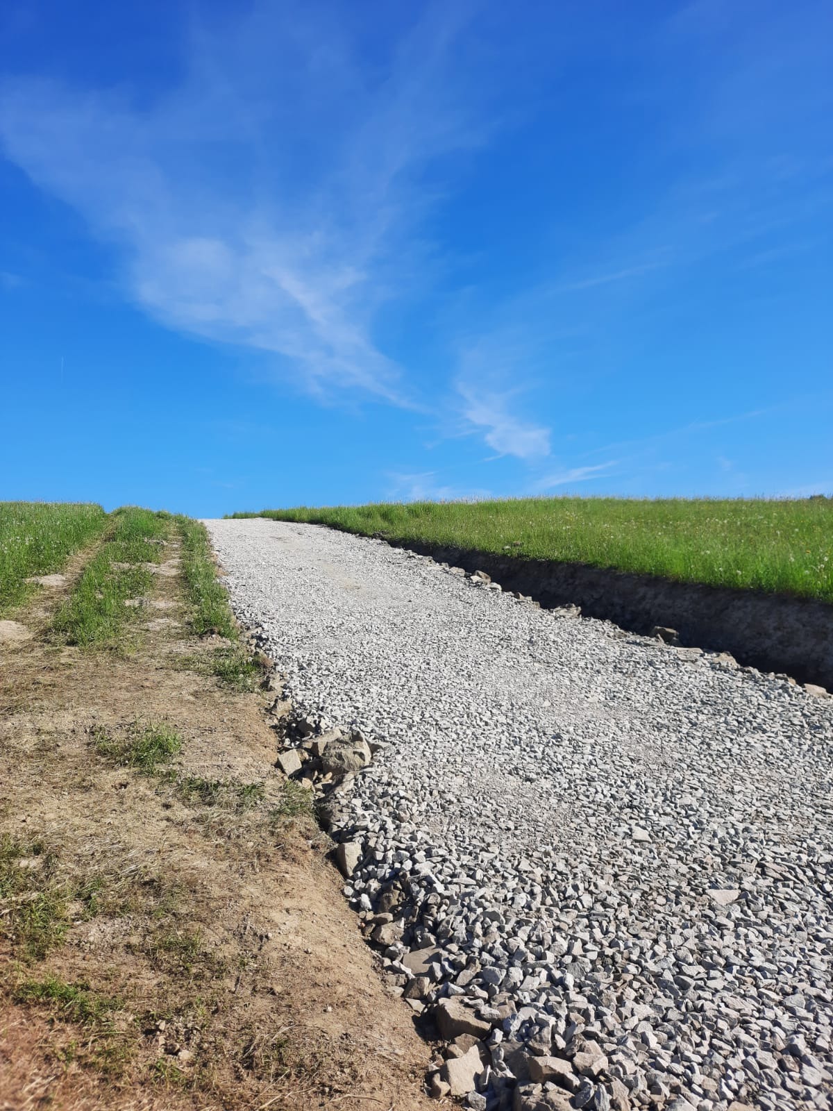 Ścieżka wysypana drobnym kruszywem na nierównym terenie, widok z dołu pod kątem, błękitne niebo z delikatnymi chmurami w tle.