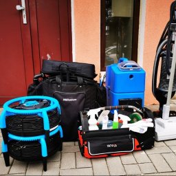 ibezplam - Zestaw sprzętu do czyszczenia: wentylator z niebieskimi elementami, torba Millenium, niebieski odkurzacz piorący, odkurzacz pionowy i torba YATO z detergentami, na tle drzwi i okna.