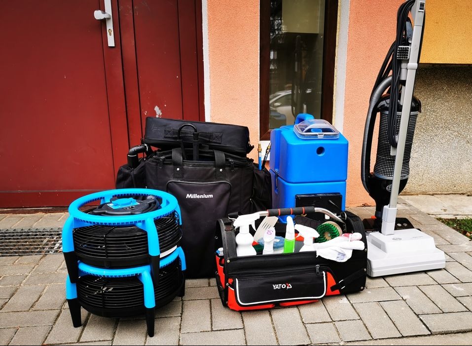 Zestaw sprzętu do czyszczenia: wentylator z niebieskimi elementami, torba Millenium, niebieski odkurzacz piorący, odkurzacz pionowy i torba YATO z detergentami, na tle drzwi i okna.