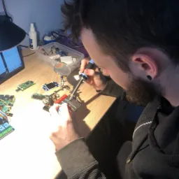 Mężczyzna z brodą lutuje element elektroniczny na płytce prototypowej przy użyciu lutownicy, wspomagając się lupą. Na biurku widoczne inne płytki, laptop i pojemnik z elektroniką.