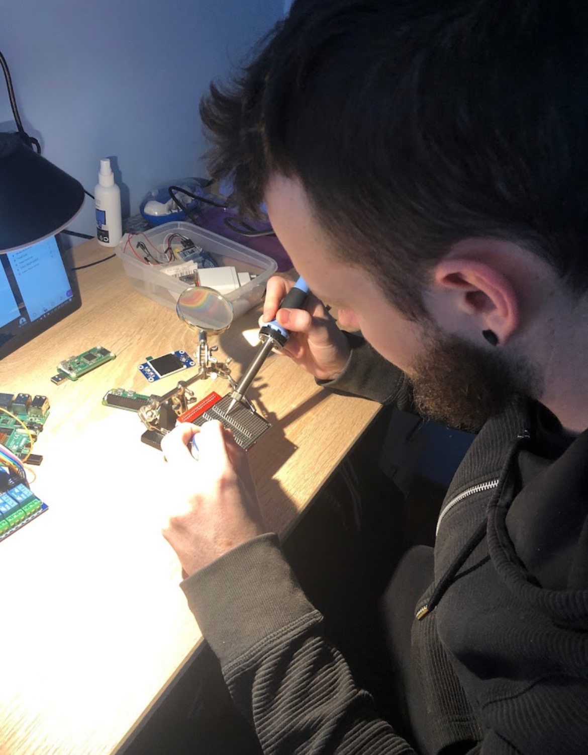 Mężczyzna z brodą lutuje element elektroniczny na płytce prototypowej przy użyciu lutownicy, wspomagając się lupą. Na biurku widoczne inne płytki, laptop i pojemnik z elektroniką.