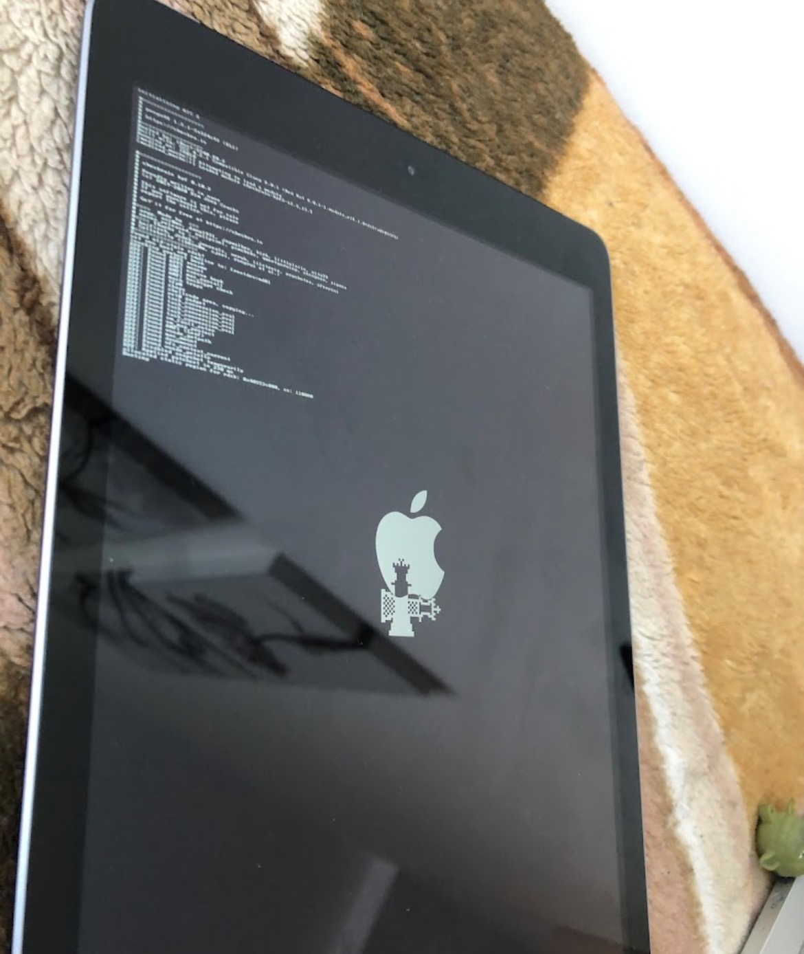 Tablet z wyświetlonym logo Apple zmodyfikowanym elementami szachowymi i tekstem w trybie debugowania, leżący na wzorzystym tle.