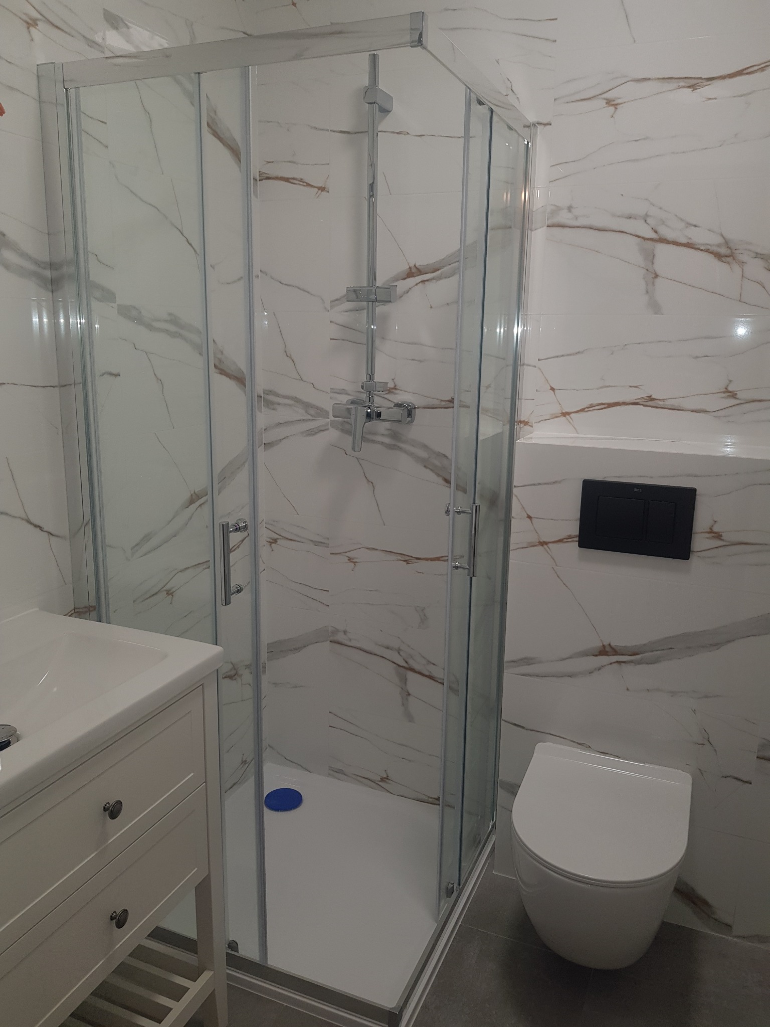 Nowoczesna łazienka z kabiną prysznicową, umywalką z szafką w stylu prowansalskim i podwieszaną toaletą, wykończona płytkami imitującymi marmur z brązowymi żyłami.