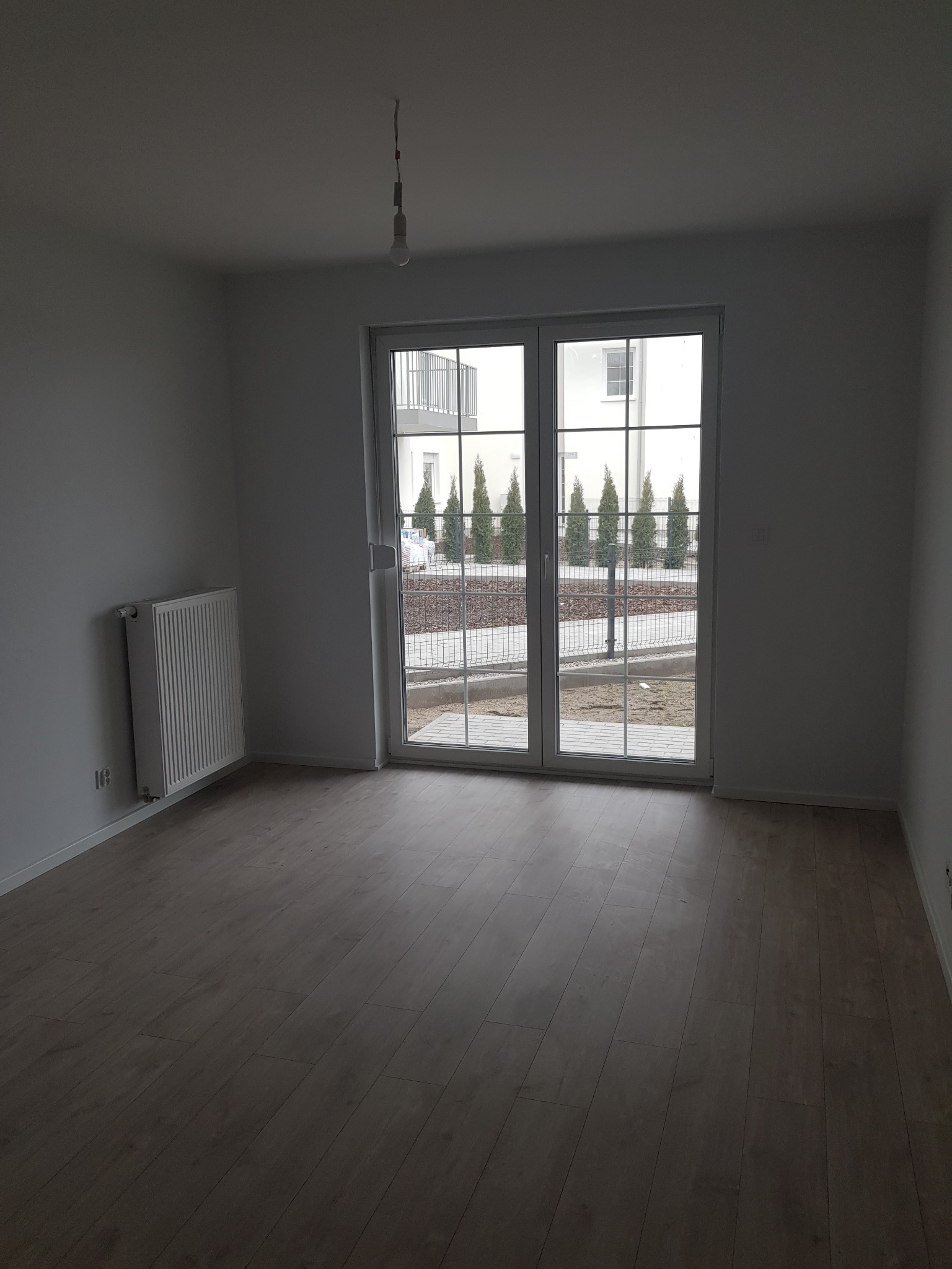 Pusty pokój z drewnianą podłogą, białymi ścianami, grzejnikiem i dużym oknem balkonowym z widokiem na podwórze z tujami.