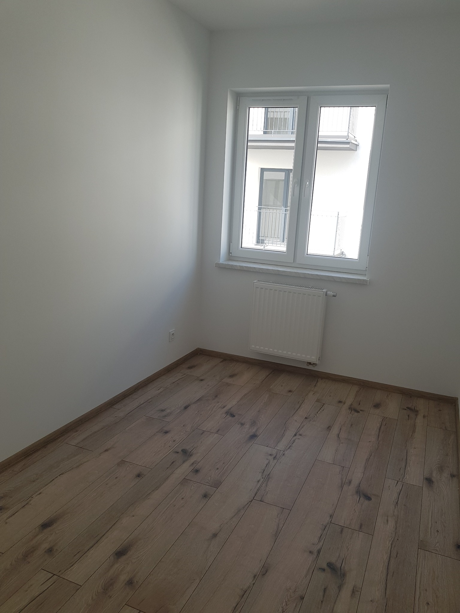 Pusty pokój po remoncie z panelami podłogowymi w odcieniach brązu, białymi ścianami i oknem z widokiem na balkon.