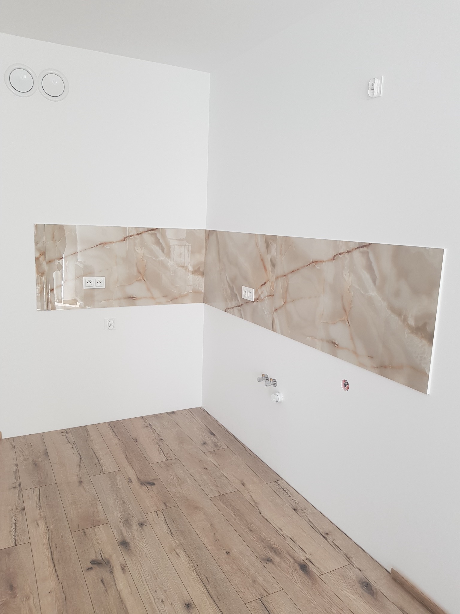 Fragment kuchni z panelami ściennymi imitującymi marmur, widoczne gniazdka elektryczne i instalacja wodno-kanalizacyjna, podłoga wyłożona panelami drewnopodobnymi.