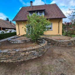 Dariusz Sek - Kaskadowy ogródek z kamiennymi murkami oporowymi przed żółtym domem z czerwoną dachówką, świeżo usypana ziemia przygotowana do nasadzeń, widoczne ślady prac ziemnych.