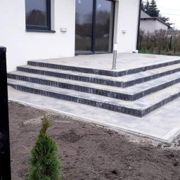 Dariusz Sek - Nowoczesne schody zewnętrzne z szarej kostki brukowej prowadzące do domu z dużym oknem tarasowym, widoczne młode tuje w gruncie i elementy oświetlenia.