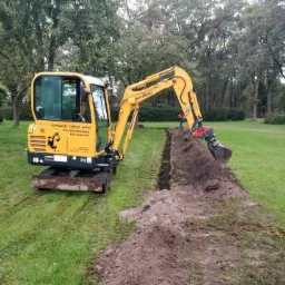 Żółta minikoparka Hyundai wykonuje wąski wykop w trawiastym terenie, z widocznym stosem ziemi po jednej stronie wykopu i drzewami w tle.
