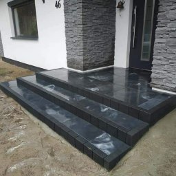 Dariusz Sek - Nowoczesne schody wejściowe z ciemnego kamienia, wykończone kostką, prowadzące do domu z elewacją w kolorze białym i szarym kamieniem dekoracyjnym, widoczne błoto na podłożu.