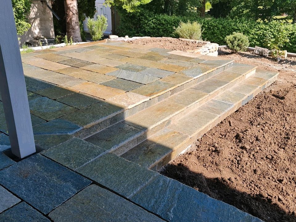 Kamienny taras z trzema stopniami schodów, wykonany z płytek o zróżnicowanej kolorystyce odcieni żółci, zieleni i szarości, częściowo otoczony ziemią, z widocznym słupem konstrukcyjnym i zielenią...