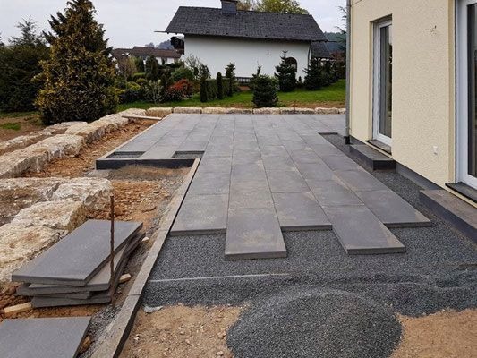 Montaż tarasu z płyt kamiennych na podsypce żwirowej, widoczne niedokończone fragmenty i zapas płyt obok kamiennego murku oporowego.