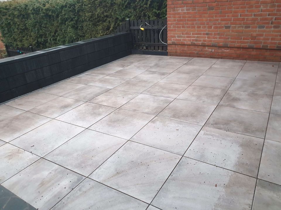 Wyłożony jasnoszarymi płytami taras przylegający do ściany z czerwonej cegły i czarnego murku z roślinnością w tle.