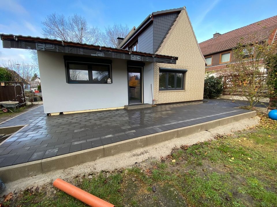 Nowo ułożony taras z ciemnej kostki brukowej przylegający do domu z cegły i szarego tynku, widoczny fragment ogrodu i rura PCV.