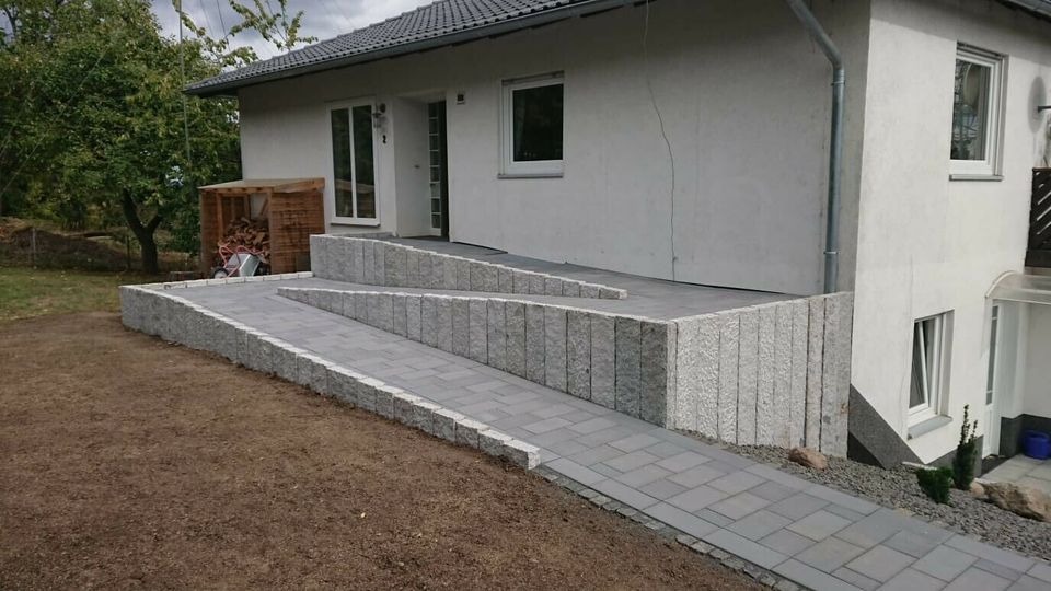 Wykonana z szarej kostki granitowej serpentynowa ścieżka prowadząca do domu w Gdańsku, otoczona niskim murem z pionowo ułożonych kamieni, widoczna zieleń trawnika i drzew.