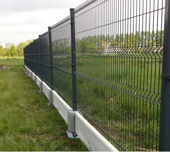 Długi, ciemnoszary panelowy płot z podmurówką z prefabrykatów betonowych na zewnątrz, widok z boku na tle zielonej trawy i drzew.