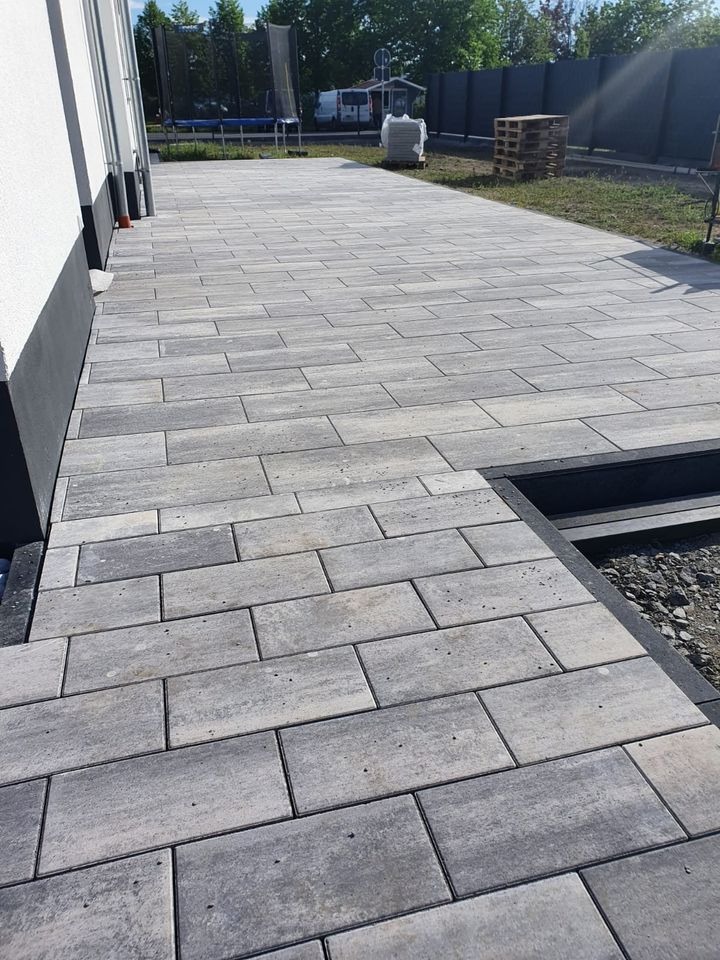 Nowo ułożony, szary chodnik z prostokątnej kostki brukowej, z widocznym obrzeżem i przygotowanym podłożem, prowadzący w kierunku trampoliny i palet w tle.