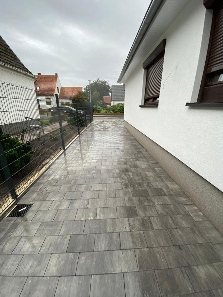 Ułożona szara kostka brukowa prowadząca wzdłuż białego domu z brązowymi okiennicami, widoczna czarna metalowa siatka ogrodzeniowa i mokra nawierzchnia po deszczu.