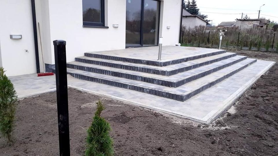 Nowoczesne schody zewnętrzne z szarej kostki brukowej prowadzące do domu z dużym oknem tarasowym, widoczne młode tuje w gruncie i elementy oświetlenia.