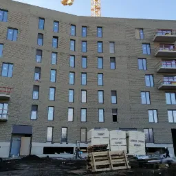 Wielopiętrowy budynek w trakcie budowy, widoczne rusztowania, dźwig, mini koparka oraz materiały budowlane na placu budowy.