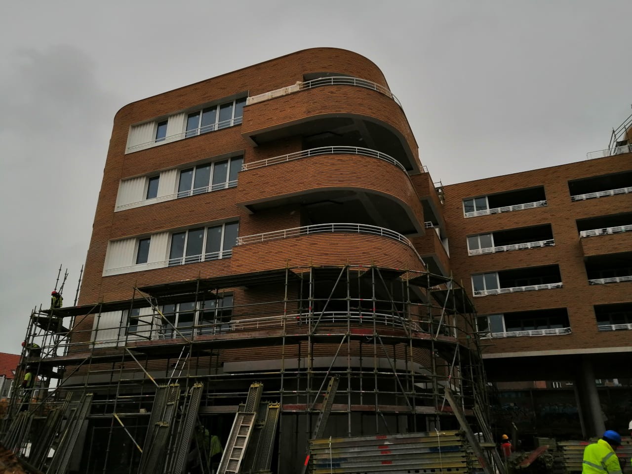 Budynek wielorodzinny w trakcie budowy, z zaokrąglonymi balkonami i ceglaną elewacją, otoczony rusztowaniami i elementami konstrukcyjnymi.