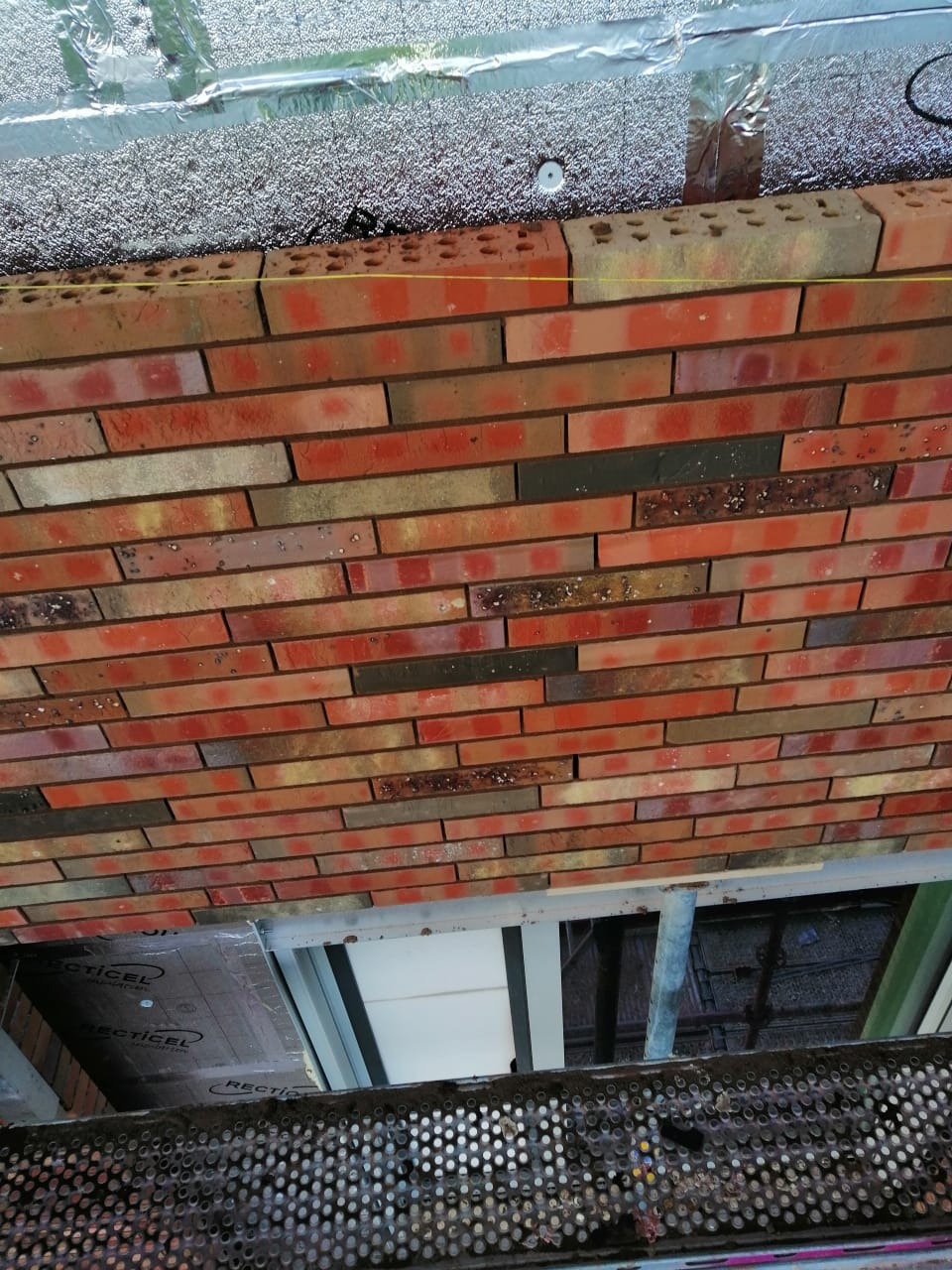 Ceglany mur widziany z rusztowania, z widocznym sznurkiem murarskim wyznaczającym poziom, oraz izolacją termiczną nad murem.