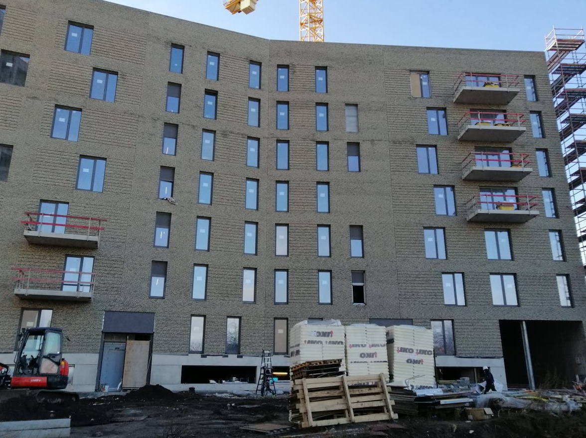 Wielopiętrowy budynek w trakcie budowy, widoczne rusztowania, dźwig, mini koparka oraz materiały budowlane na placu budowy.