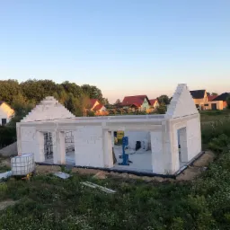 Wznoszenie ścian domu z białych bloczków Ytong, widoczna piła taśmowa do cięcia bloczków i zbiornik na wodę, w tle zabudowa jednorodzinna i drzewa.