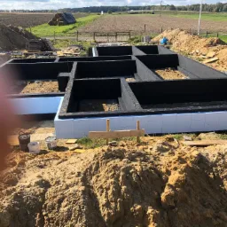 Wykopane fundamenty z izolacją termiczną, przygotowane do dalszych prac budowlanych na tle krajobrazu wiejskiego z belami siana.