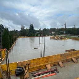 Denis Los MAXBAU - Zalany beton na fundamencie z wystającymi prętami zbrojeniowymi i żółtym szalunkiem. Widoczne narzędzia budowlane i otoczenie z zielenią. Pochmurne niebo w tle.