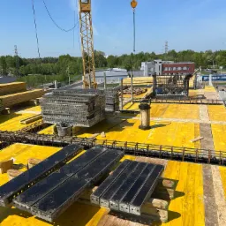 Ż&oacute;łty szalunek stropowy z ułożonym zbrojeniem i stosem metalowych element&oacute;w, widok z g&oacute;ry na budowę z dźwigiem w tle, słoneczny dzień.