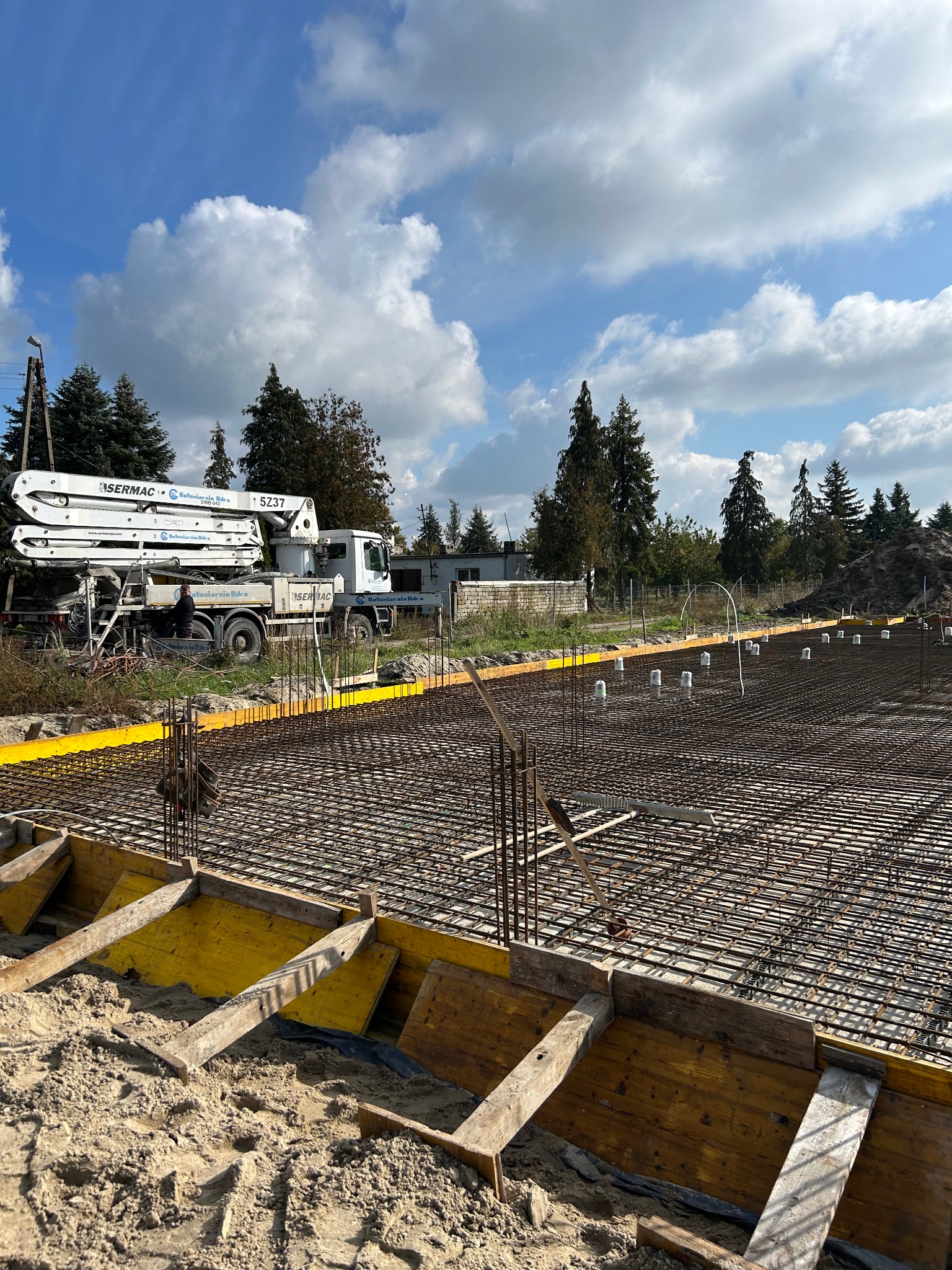 Zbrojenie fundamentów pod budowę domu jednorodzinnego, widok z góry na siatkę zbrojeniową i szalunki, w tle pompa do betonu i błękitne niebo.