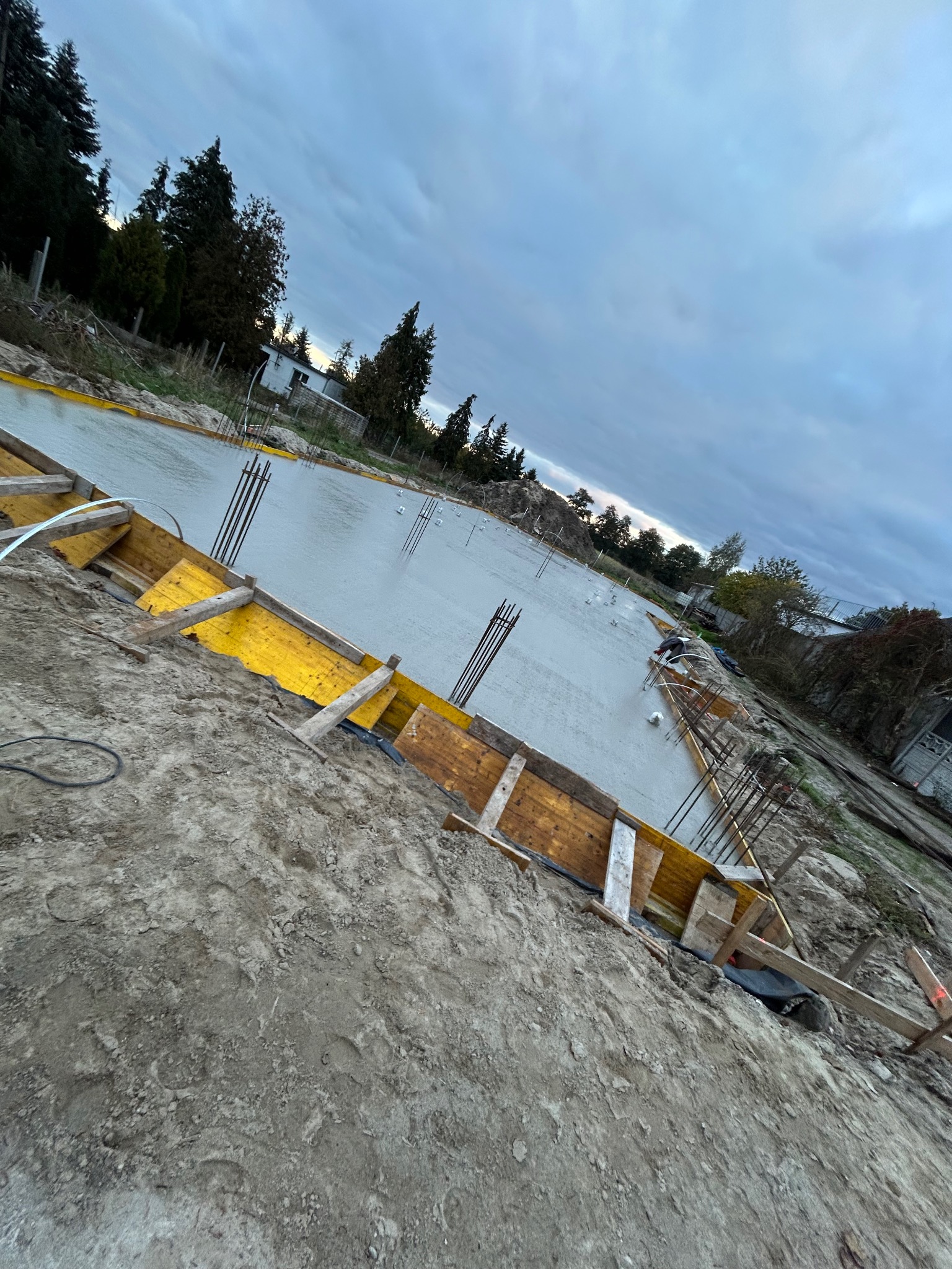 Zalany beton w szalunku z żółtą folią i wystającymi prętami zbrojeniowymi, widok z góry pod kątem, pochmurne niebo w tle.