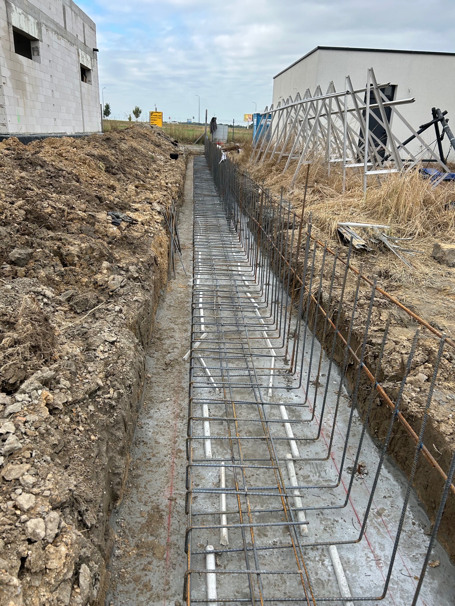 Zbrojenie ławy fundamentowej w wykopie, widoczne białe rury instalacyjne. W tle budynek w trakcie budowy i konstrukcja wsporcza.