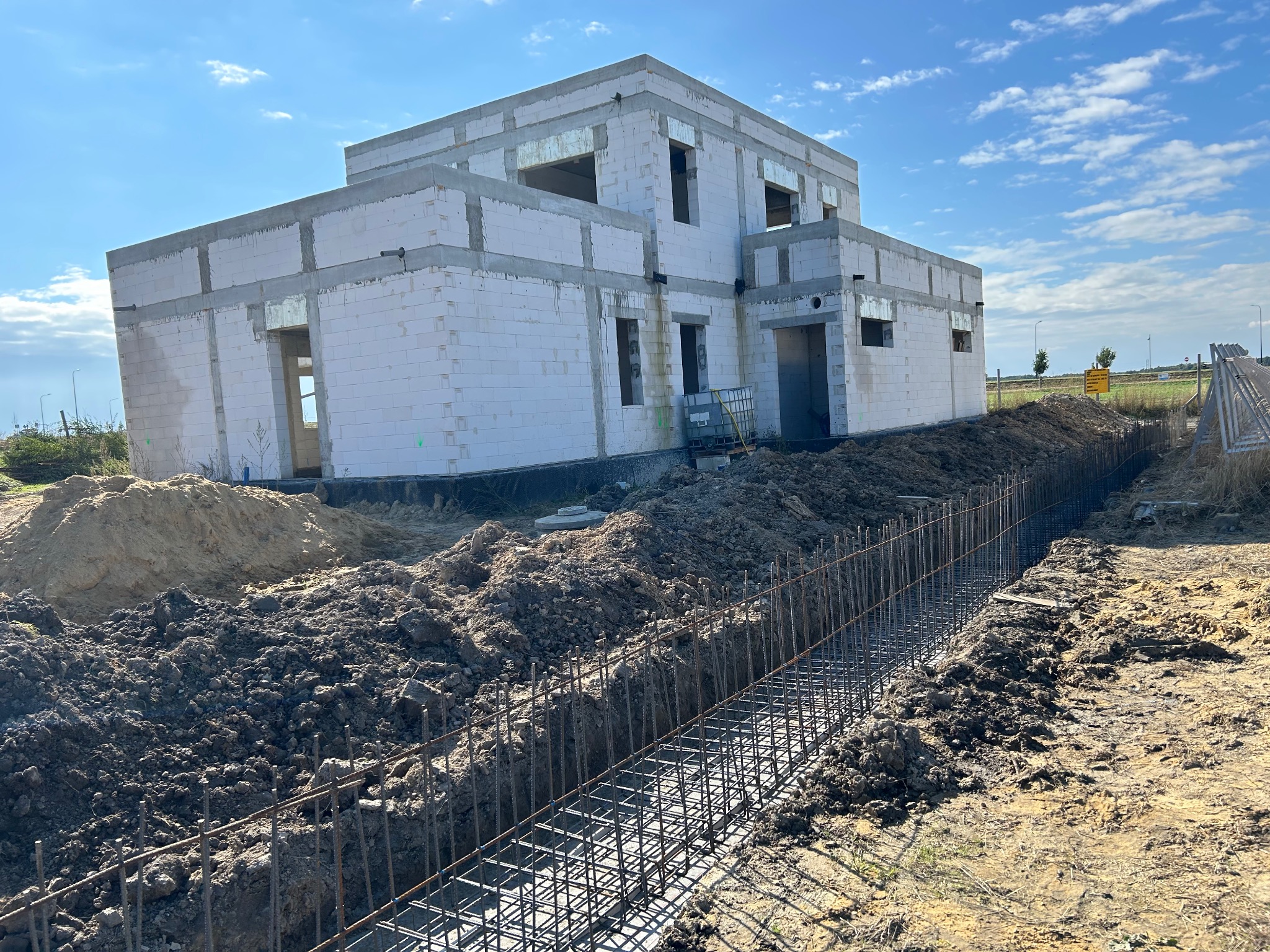 Dwukondygnacyjny dom w stanie surowym otwartym z białego betonu komórkowego, widoczny wykop zbrojony pod fundamenty. Słoneczny dzień, błękitne niebo.