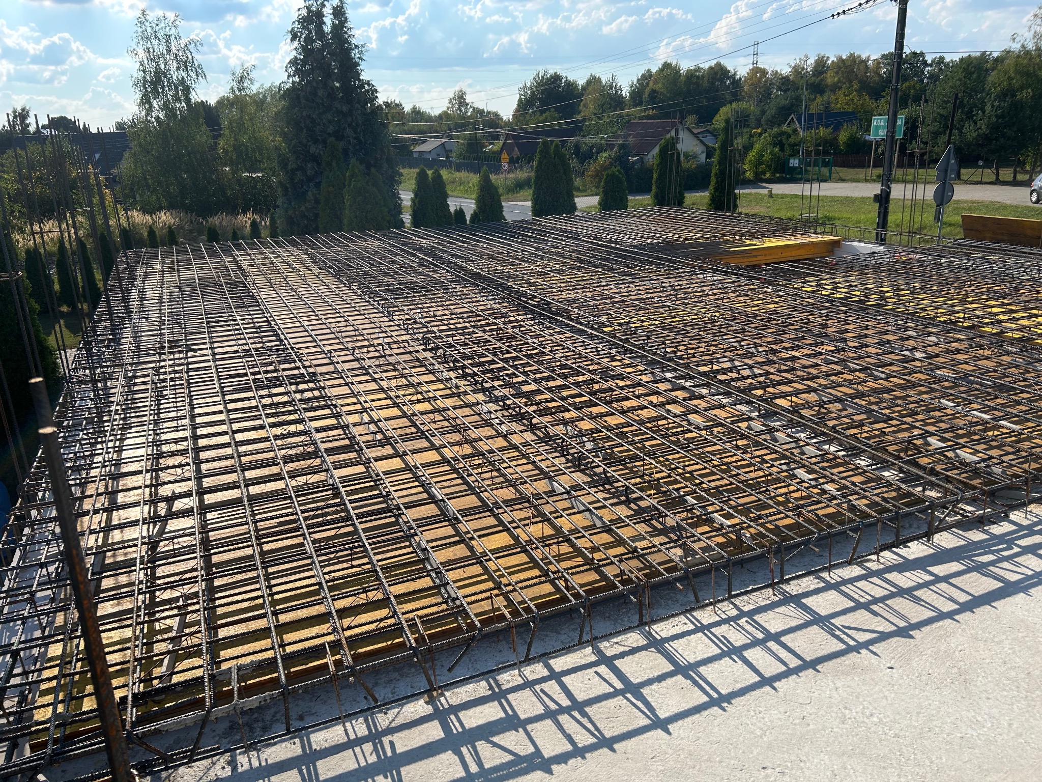 Siatka zbrojeniowa na żółtym szalunku, przygotowana do zalania betonem. Widok z góry na budowę z zielonym tłem drzew i niebieskim niebem.