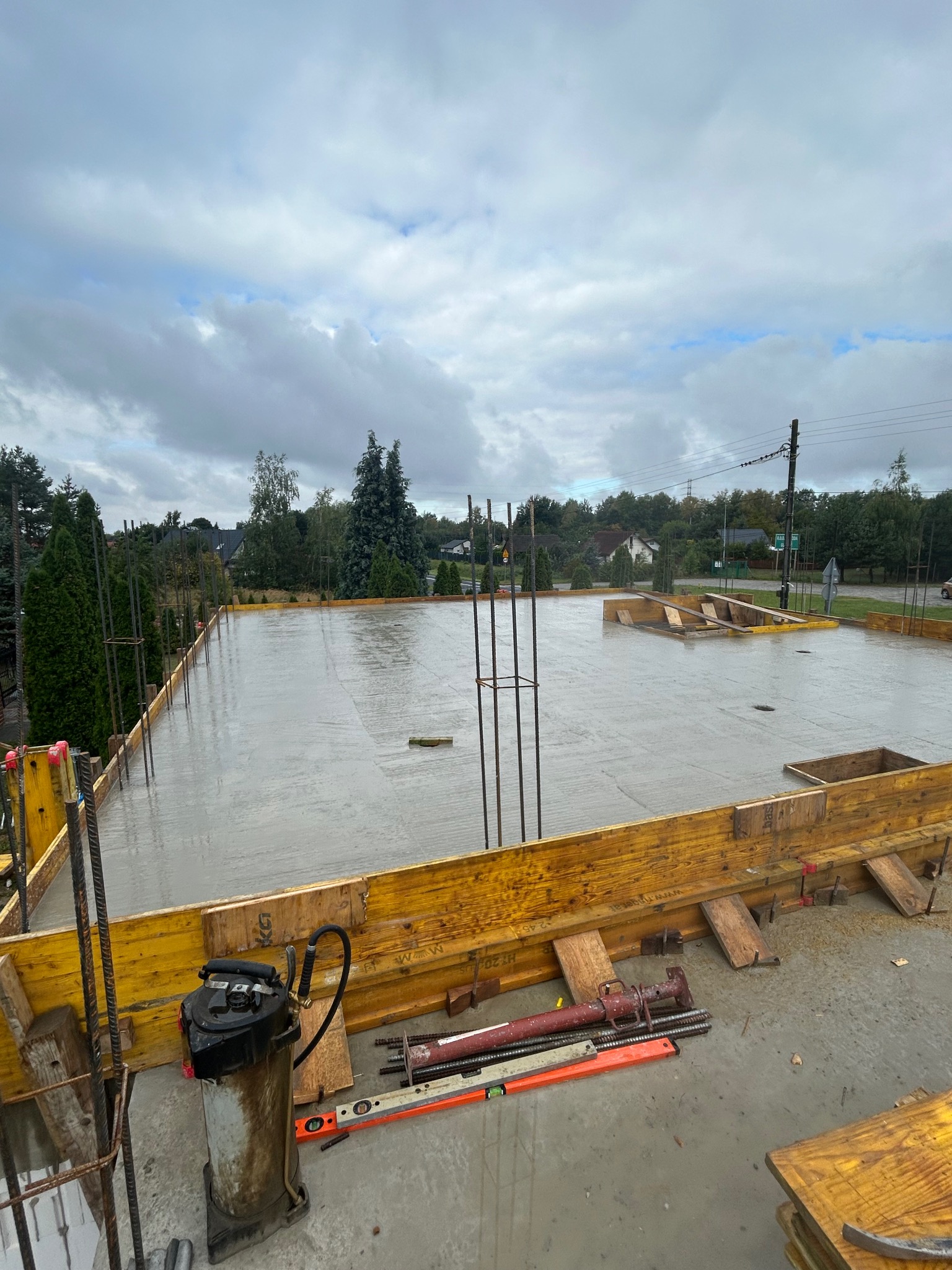 Zalany beton na fundamencie z wystającymi prętami zbrojeniowymi i żółtym szalunkiem. Widoczne narzędzia budowlane i otoczenie z zielenią. Pochmurne niebo w tle.
