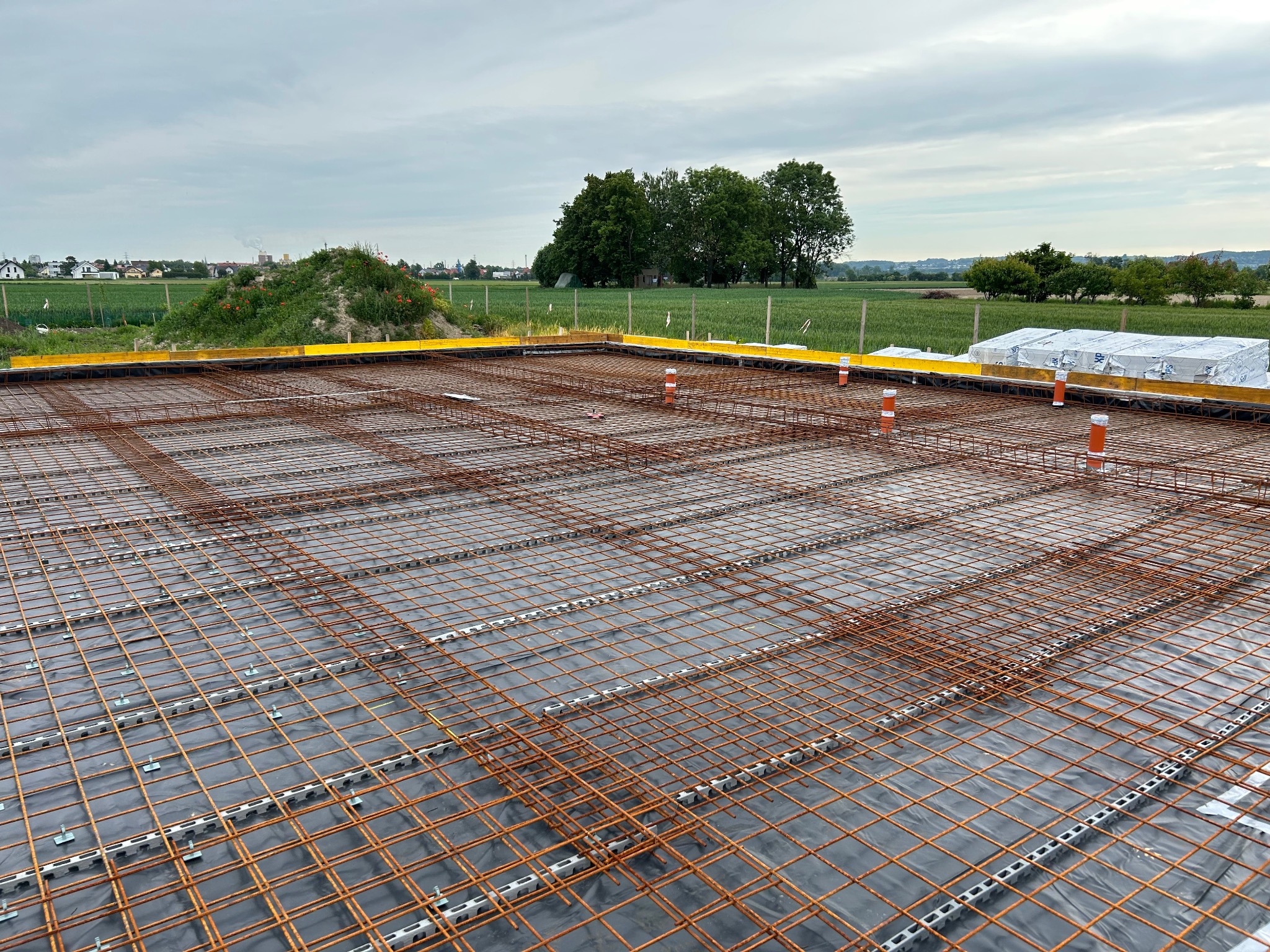 Zbrojenie fundamentów na czarnej folii, widoczne pomarańczowe rury i żółte zabezpieczenia krawędzi. W tle zielone pola i zabudowania.