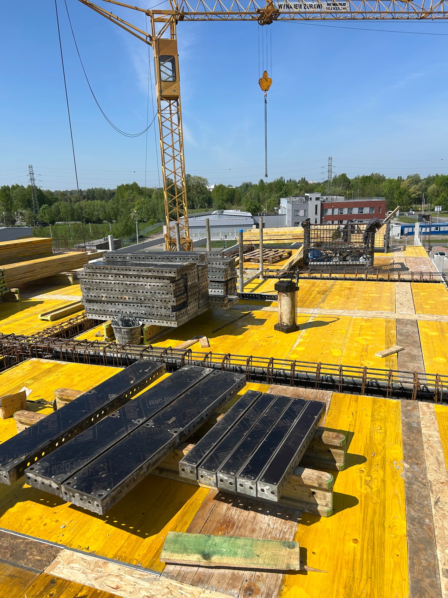 Żółty szalunek stropowy z ułożonym zbrojeniem i stosem metalowych elementów, widok z góry na budowę z dźwigiem w tle, słoneczny dzień.