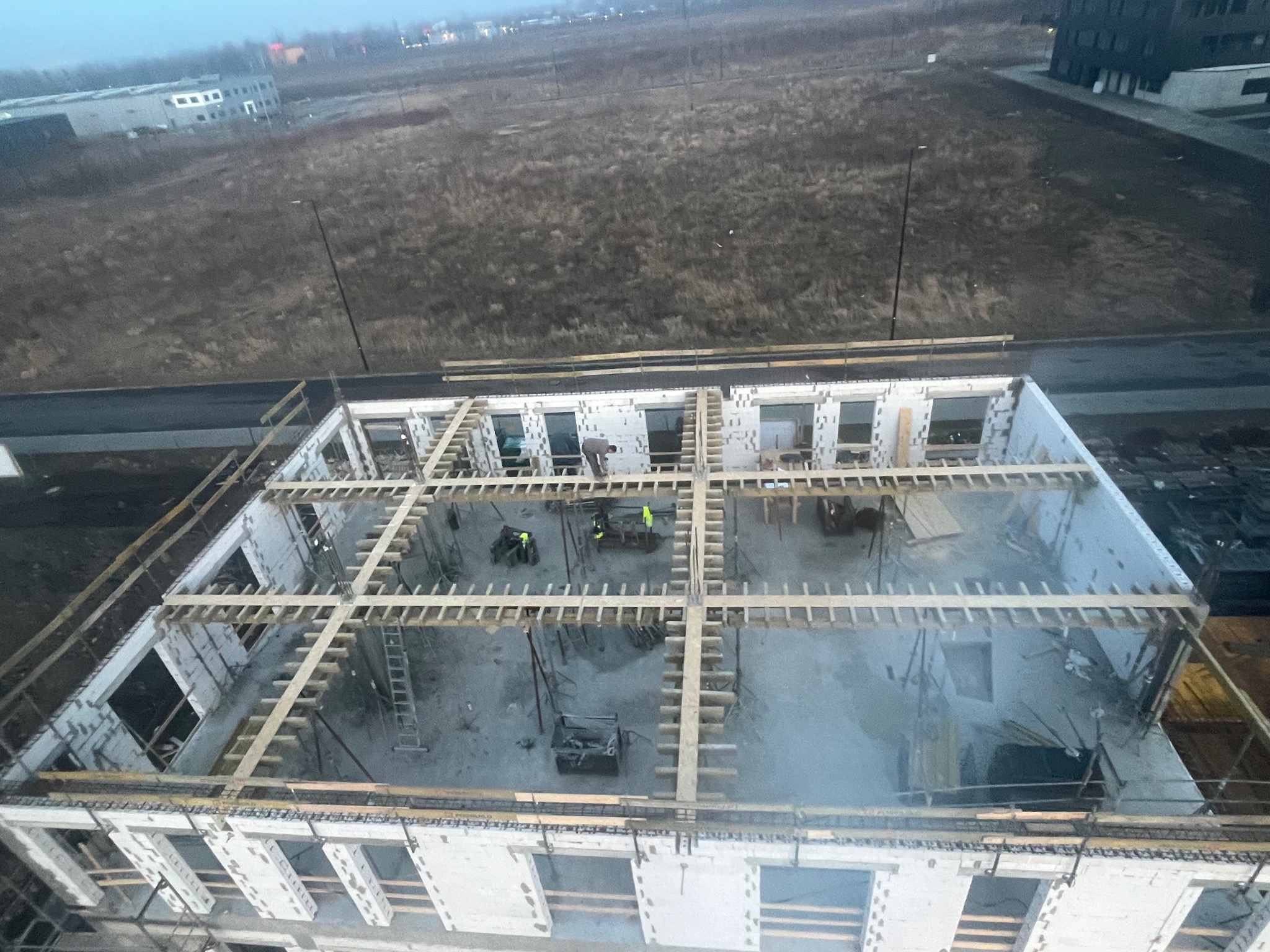 Budowa domu z betonu komórkowego: widok z góry na wznoszone ściany i drewniane szalunki stropu. Dwóch pracowników w żółtych kamizelkach na budowie.