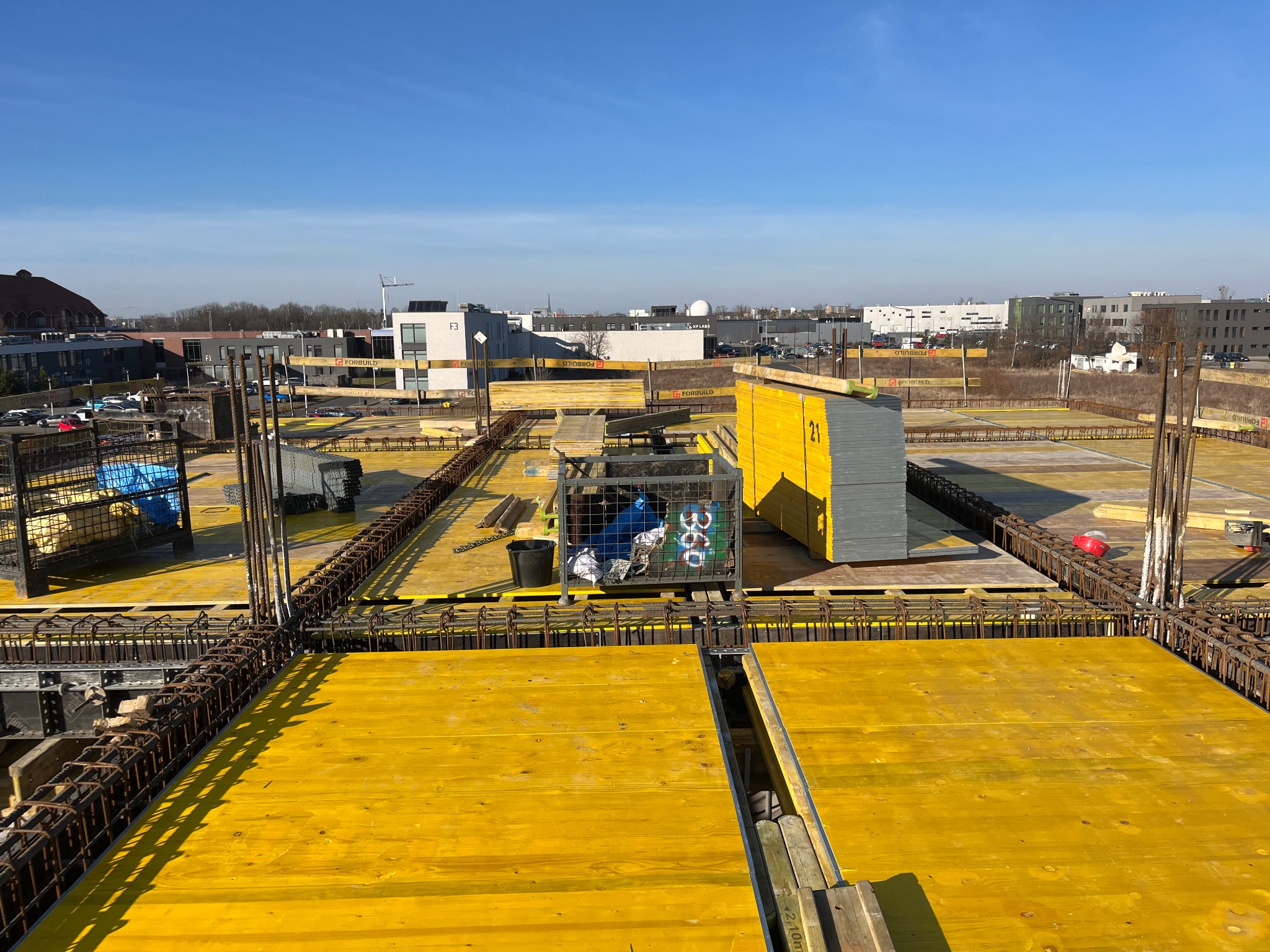 Żółte szalunki i zbrojenia na budowie, widok z góry w słoneczny dzień. W tle budynki miejskie i błękitne niebo. Elementy konstrukcyjne w trakcie realizacji.